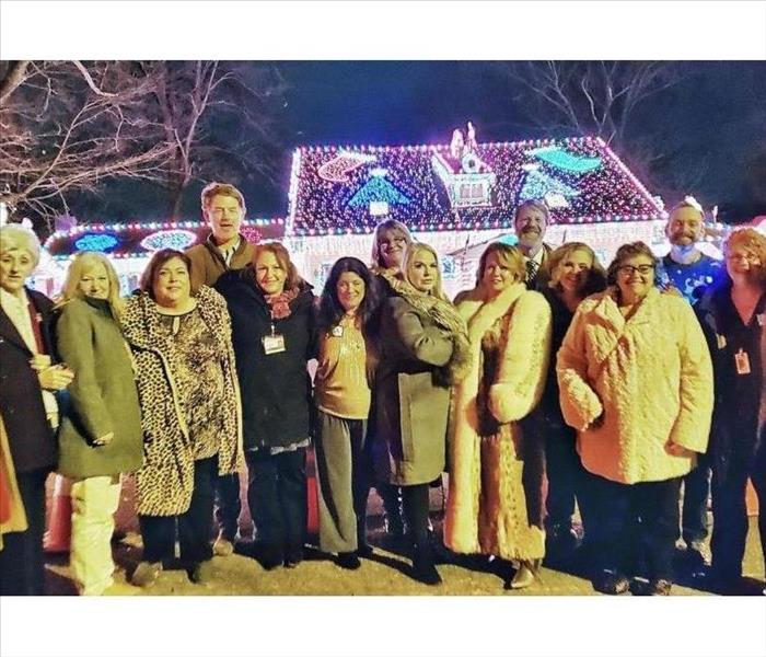 large group of men and women standing in front of a house decorated with Christmas lights